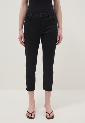 Mujer con jeans negros ajustados y cortos y sandalias negras de dedo, de pie frente a un fondo claro y liso.