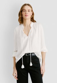 Blusa leggera e bianca con colletto arricciato e scollatura a V. Presenta cordini con nappine. Indossata sopra jeans neri, con vestibilità rilassata.