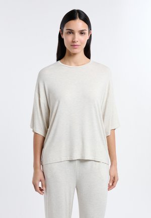 Donna con lunghi capelli scuri che indossa una maglietta beige chiaro a maniche corte e pantaloni abbinati, in piedi davanti a uno sfondo bianco uniforme.