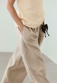 Person trägt beige weite Hosen und ein cremefarbenes gestricktes ärmelloses Top, mit Händen in den Taschen und einem schwarzen gehäkelten Accessoire.