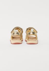 Złote metaliczne sandały na paskach z tyłu, z brązową wkładką, zaokrąglonym kształtem i gumową podeszwą z czerwonymi akcentami. Otwarta konstrukcja zapewniająca wentylację.