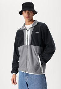 Fleece zip-up jacket in black and gray with a white zipper, two side pockets, and a high collar. Worn with loose blue denim pants and a black bucket hat.