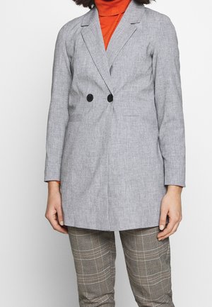 Frau trägt hellgrauen zweireihigen Blazer, orange Rollkragenpullover und karierte Hose und steht vor einem einfarbigen Hintergrund.