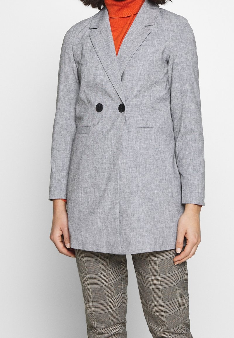 Femme portant un blazer gris clair à double boutonnage, un col roulé orange et un pantalon à carreaux, debout devant un fond uni.