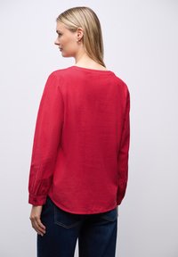 Camisa roja de manga larga hecha de tela ligera con cuello redondo y puños abotonados, combinada con pantalones vaqueros azules. Textura suave y corte relajado.