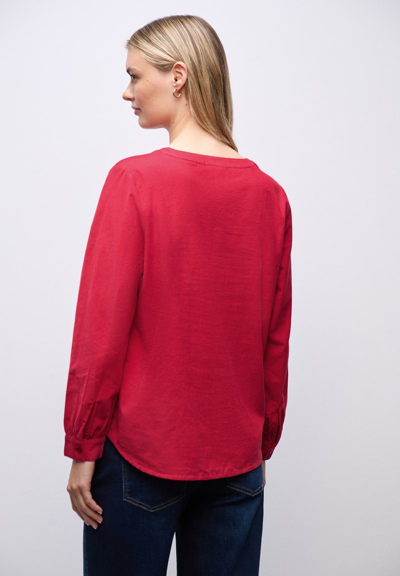 Camisa roja de manga larga hecha de tela ligera con cuello redondo y puños abotonados, combinada con pantalones vaqueros azules. Textura suave y corte relajado.