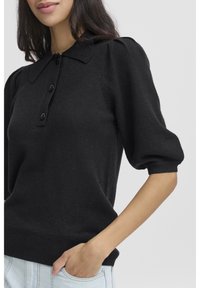 Juodas megztinis su apykakle, sagomis užsegamas priekyje ir pūstas trumpomis rankovėmis; turi ribotą apačią ir minkštą tekstūrą.