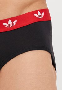 Fekete pamut alsónadrág piros gumírozott derékkal, fehér Adidas trefoil logóval díszítve és sima textúrával.