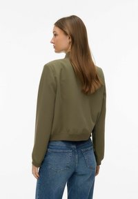 Veste bomber vert olive avec un ourlet et des poignets côtelés, associée à un jean bleu taille haute. Tissu lisse, design minimaliste.