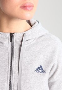 Felpa con zip grigia chiara in morbido tessuto, con cappuccio regolabile e logo Adidas blu navy sul lato sinistro del petto.