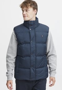 Temno moderna puhovka z visokim ovratnikom, gumbi in logotipom. Nosi se preko svetlo sive majice z dolgimi rokavi. Gladek, sijoč material.