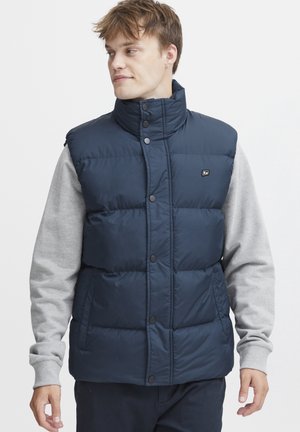 Navy blue puffy vest with high collar, button closures, and a logo. Worn over a light gray long-sleeve shirt. Smooth, shiny material.