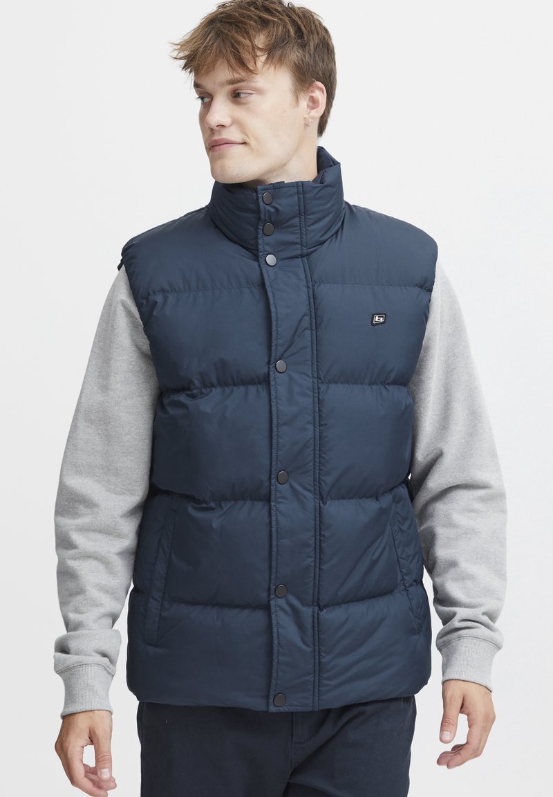 Temno moderna puhovka z visokim ovratnikom, gumbi in logotipom. Nosi se preko svetlo sive majice z dolgimi rokavi. Gladek, sijoč material.