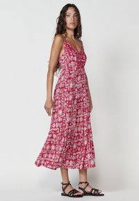 Rotes Kleid mit weißen geometrischen und floralen Mustern, ärmelloses Design, taillierte Taille und ausgestellter Rock. Kombiniert mit schwarzen Riemchensandalen.