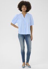 Camicia azzurra a maniche corte con bottoni, colletto strutturato e leggeri arricciamenti, abbinata a jeans aderenti blu sbiaditi e tacchi slingback blu.