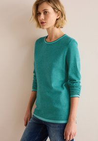 Teal stickad tröja med rund halsringning och texturerat tyg, med kontrasterande ljus teal ribbade manschetter och nederkant. Sittande silhuett.