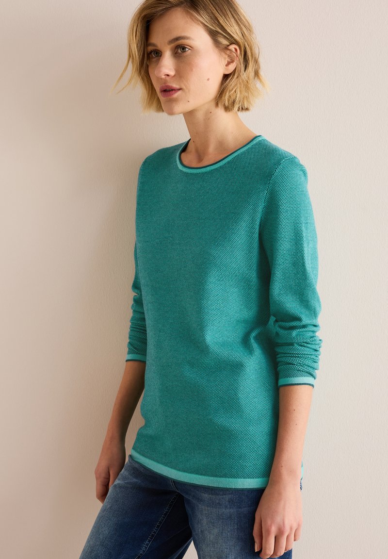 Teal stickad tröja med rund halsringning och texturerat tyg, med kontrasterande ljus teal ribbade manschetter och nederkant. Sittande silhuett.