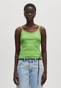 Grünes geripptes Tanktop mit breiten Trägern, kombiniert mit hellblauen, hoch taillierten Jeans, die am Bund ein schwarzes Kordelzugdetail aufweisen.
