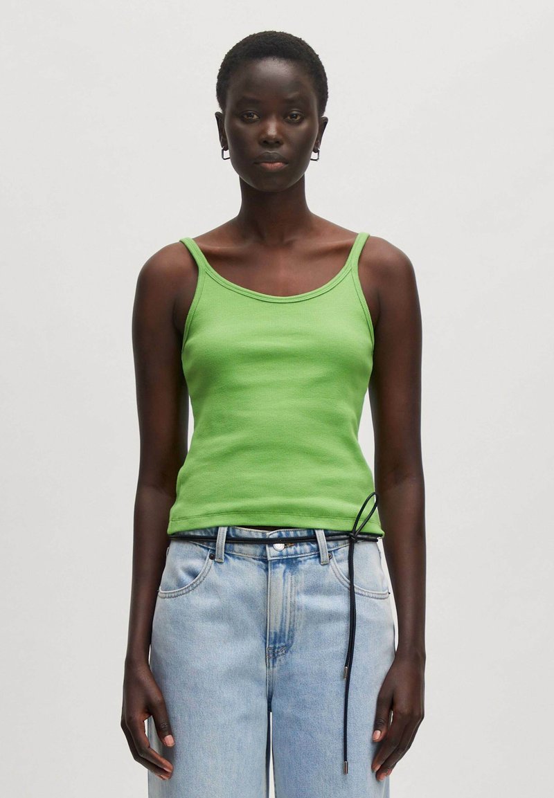 Grünes geripptes Tanktop mit breiten Trägern, kombiniert mit hellblauen, hoch taillierten Jeans, die am Bund ein schwarzes Kordelzugdetail aufweisen.