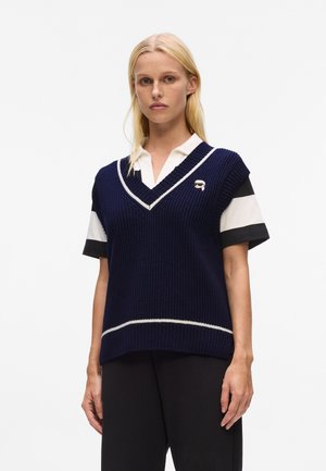 Navy knitted sweater vest with white trim and v-neck, paired with a black and white collared shirt; features close-fitting sleeves.