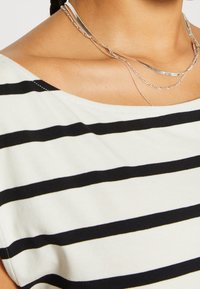 Top de rayas en crema y negro, con un escote barco, corte holgado y textura suave. Collares plateados en capas añaden detalle.
