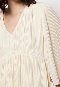 Helles beiges, gefältetes Kleid mit V-Ausschnitt, fließenden Ärmeln und weicher Textur. Der Stoff weist vertikale Falten und ein lockeres Design auf.