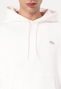 Sudadera blanca con bolsillo tipo canguro, textura acanalada, cordones ajustables y un pequeño logotipo bordado en el pecho.