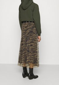 Avkortad olivgrön huvtröja som matchas med en genomskinlig, beige kjol med ett mönster av svarta tigerränder, bärs med svarta klackstövlar.