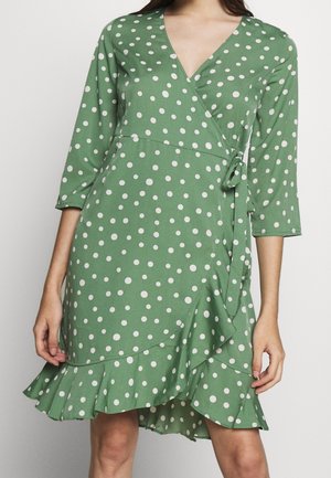 Green wrap dress with white polka dots, three-quarter sleeves, waist tie, and ruffled asymmetrical hem worn by a standing person.