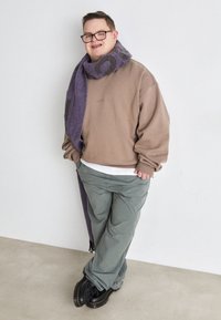 Giovane con occhiali, felpa beige, pantaloni verdi, scarpe nere e una lunga sciarpa viola, che si trova in piedi contro un muro bianco.