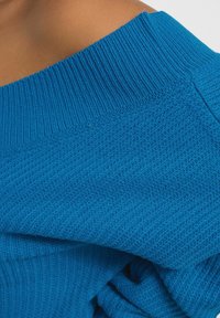 Jasnoniebieski dzianinowy wyrób z prążkowaną teksturą i odkrytymi ramionami. Charakteryzuje się luźnym krojem oraz widocznymi detalami szycia.