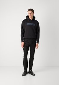 Hanorac negru cu glugă, având un logo pe față, combinat cu pantaloni cargo negri cu buzunare și pantofi negri cu șireturi. Textură de material neted.