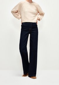 Personne portant un pull court beige clair, un jean large taille haute foncé et des chaussures marron à bout carré, debout les mains sur les hanches.