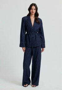 Denim blejzer z globokim V-izrezom, dolgimi rokavi in sprednjimi gumbi. Usklajen z širokimi kavbojkami, obema v temnem indigu. Zaveže se v pasu.