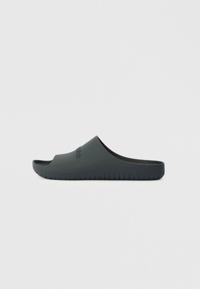 Black slide sandal made of rubber. Features a smooth surface, open toe, and a ridged outsole. Logo embossed on the side.