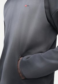 Άτομο που φοράει μακρυμάνικο πουκάμισο σκούρου γκρι χρώματος με το χέρι στην τσέπη, με μικρό κόκκινο και λευκό λογότυπο Diesel στο στήθος.