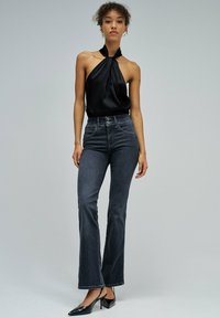 Mujer joven con top negro sin mangas halter, vaqueros oscuros de talle alto y bota ancha, y tacones negros de punta puntiaguda con tira trasera, posando frente a un fondo gris.