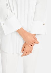 White long-sleeve shirt with pleats, featuring a tailored collar and button accents on the cuffs. Smooth, lightweight fabric.