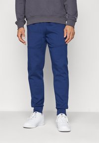 Blaue Jogginghosen aus weichem Stoff mit elastischem Bund, schmal zulaufenden Beinen und zwei vorderen Taschen. Kombiniert mit weißen Turnschuhen.
