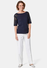 Marinebluse mit kurzen Spitzenärmeln und Verzierungen, kombiniert mit weißen Hosen und hellgrauen Slippern. Glatte Stofftextur.