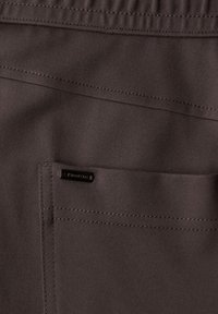 Primer plano de un bolsillo de tela marrón con costuras visibles y una pequeña etiqueta de metal negra de la marca colocada cerca del borde del bolsillo.