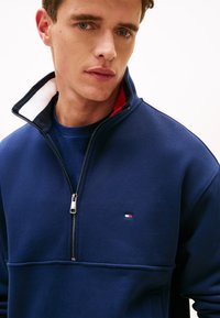 Σκούρο μπλε φούτερ με φερμουάρ και ψηλό γιακά, με κόκκινη και λευκή εσωτερική επένδυση και μικρό λογότυπο στο στήθος. Ομαλή υφή υφάσματος.