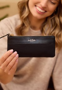 Musta lukuga rahakott, mis on valmistatud siledast nahast ja kangast, millel on kuldne logo aksessuaar ja randmepael mugavaks kandmiseks.