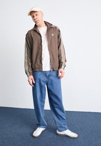 Brun zip-up jakke med stripete ermer, hvit t-skjorte, løse blå jeans og hvite joggesko på en teksturert blå teppebakgrunn.