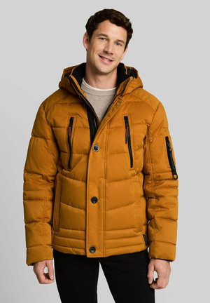 Man smiling wearing an orange quilted winter jacket with black zipper pockets and a hood, standing against a light gray background.