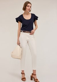 Top navy con maniche a volant, pantaloni bianchi alla caviglia con accenti a bottone laterali, cintura marrone con hardware dorato, borsa beige, sandali con tacco marrone.