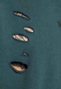 Tissu sarcelle avec cinq déchirures horizontales irrégulières révélant la peau tatouée en dessous, avec un texte noir partiel visible sur le côté droit.