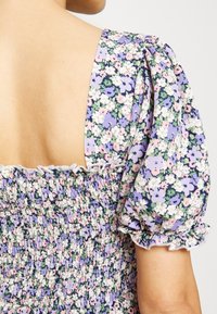 Vue de dos d'une robe fleurie avec des manches bouffantes et un tissu froncé aux couleurs pastel violet, rose, vert et blanc sur un fond clair.