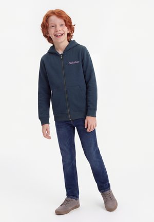 Rødhåret dreng smilende iført en mørk lynlåshættetrøje, blå jeans og grå sneakers, stående foran en ensfarvet hvid baggrund.