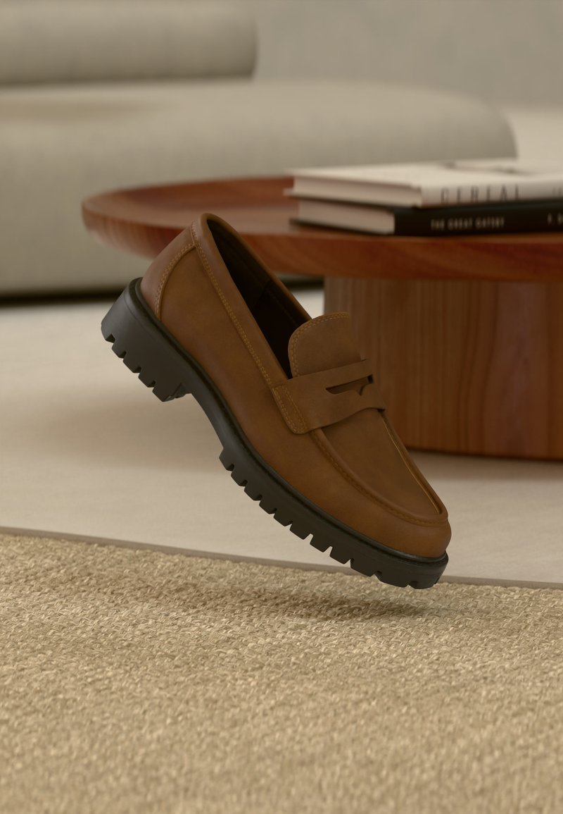 Mocassin en cuir marron avec une semelle crantée noire épaisse reposant sur un tapis beige près d'une table basse en bois avec des livres empilés.